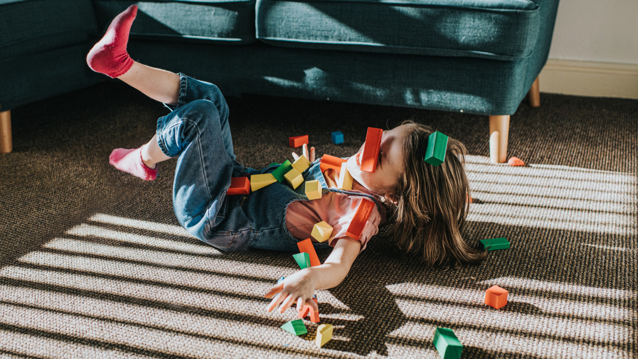 Cute little girl playfully laughs as she falls backwards. Her feet are up in the air and her hand casts a shadow on the sunny carpet. She is covered in colourful wooden blocks that scatter all around her as she bumps on the ground.