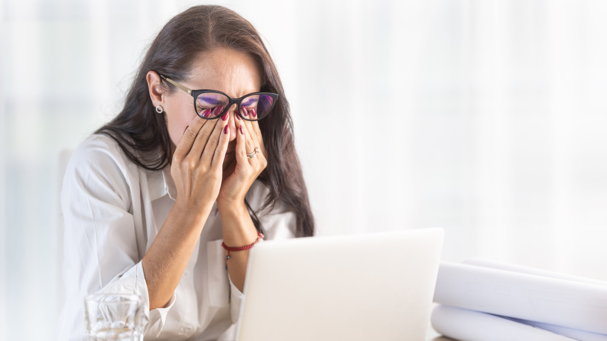 Woman in the office scratches her eyes behind the eyewear as long computer work causes headache.