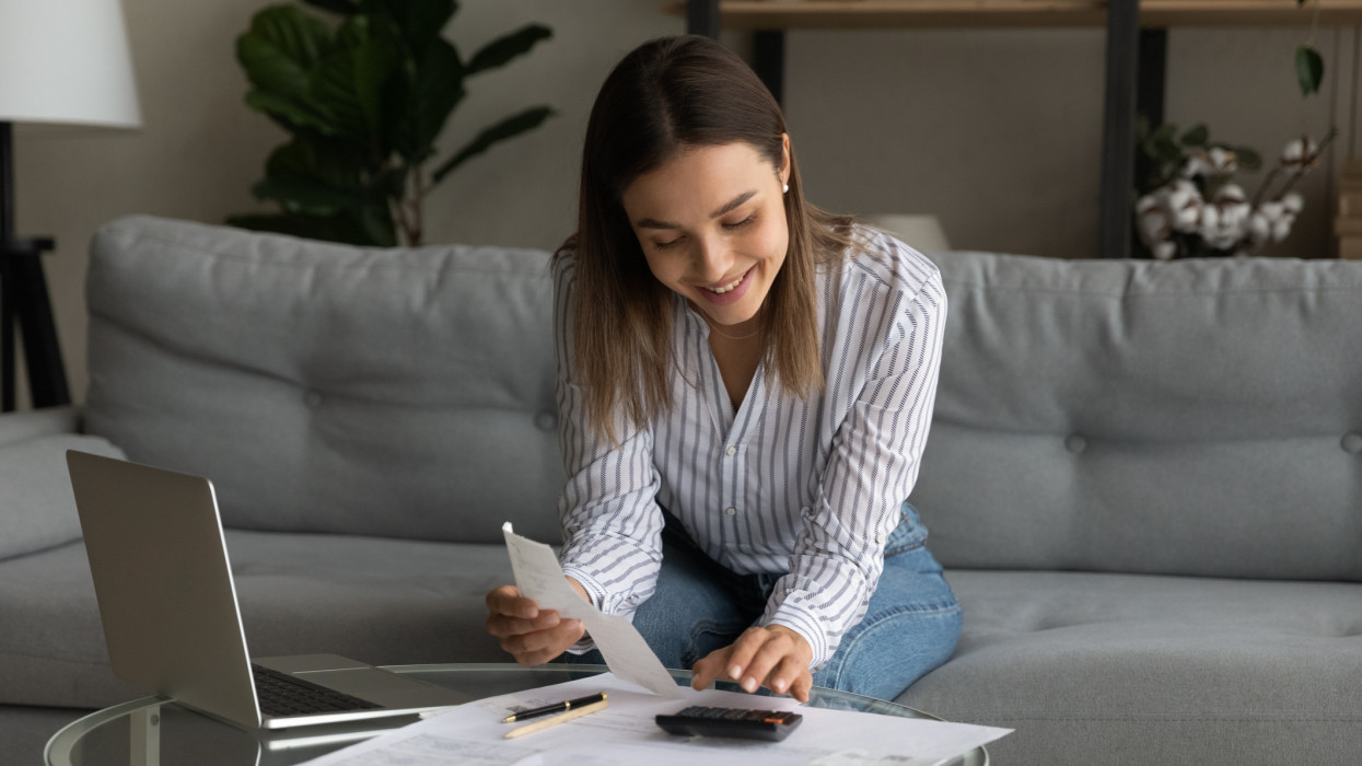 Happy young woman holding paper bills in hands, calculating expenditures, paying bills or services online in computer application, feeling satisfied with enough money, investment payments concept.