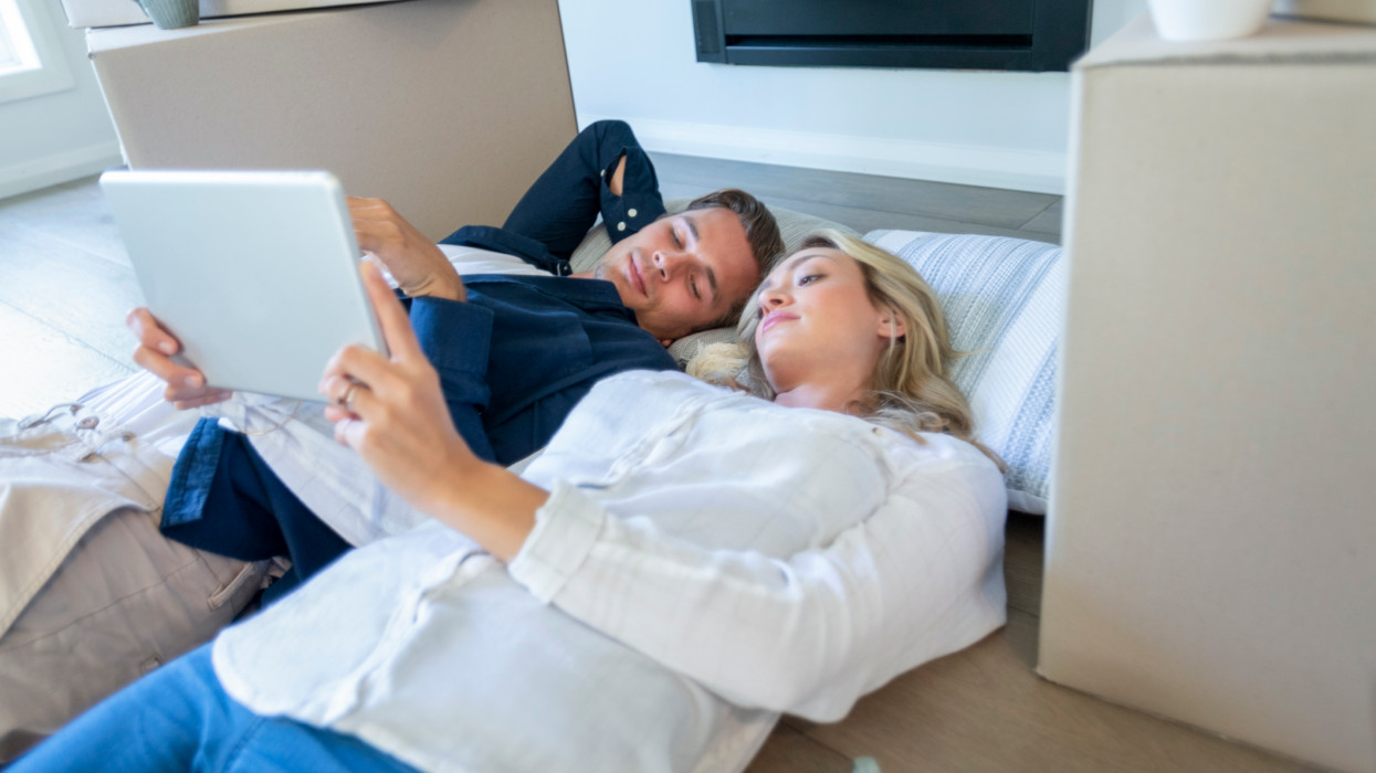 Couple resting while moving house. They could be packing or unpacking the boxes around them. They are both looking at a digital tablet. They are both looking a little worried and concerned.