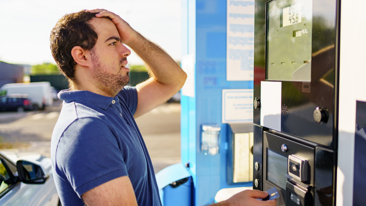 Caucasian young man surprised by high fuel prices on the gas station scoreboard
