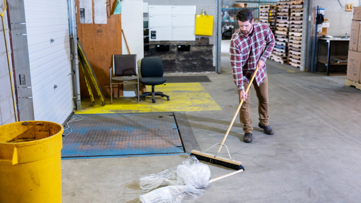 Warehouse safety topic.  A man cleaning up debris and trash at a loading dock in a warehouse to avoid slip and trip hazards.