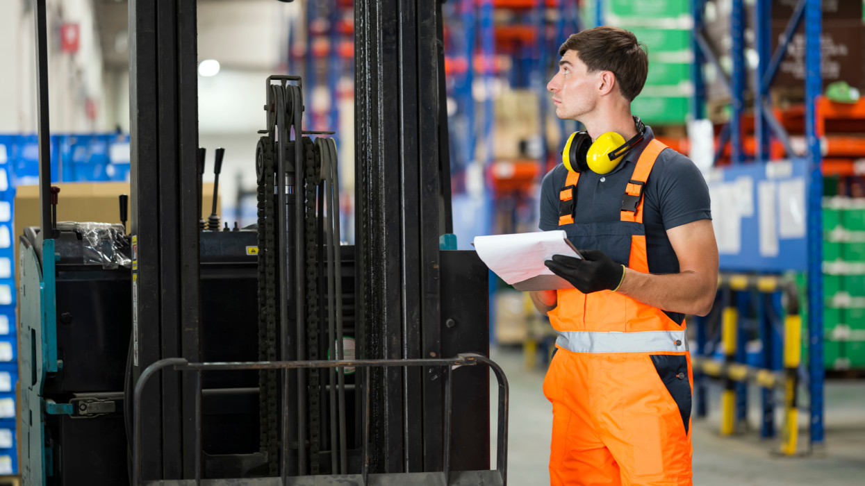 Electric Forklifts Pre-operation Checklist. Male Maintenance technician or forklift driver is checking battery restraints are functional and record checklist report at a factory warehouse.