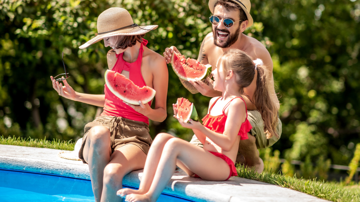 Family sitting on the edge of a pool, having fun and eating watermelon.