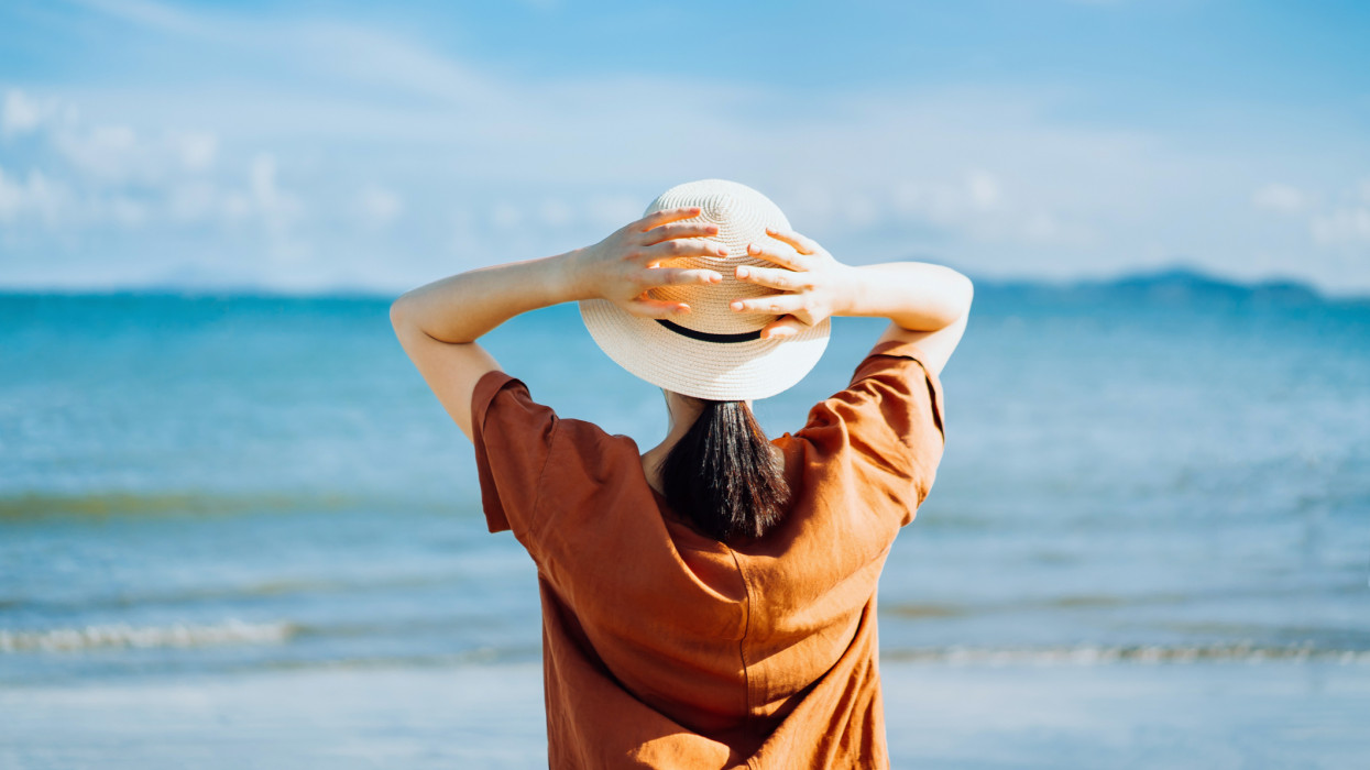Rear view of carefree young Asian woman with straw hat relaxing on the beach against blue sky on a sunny Summer day