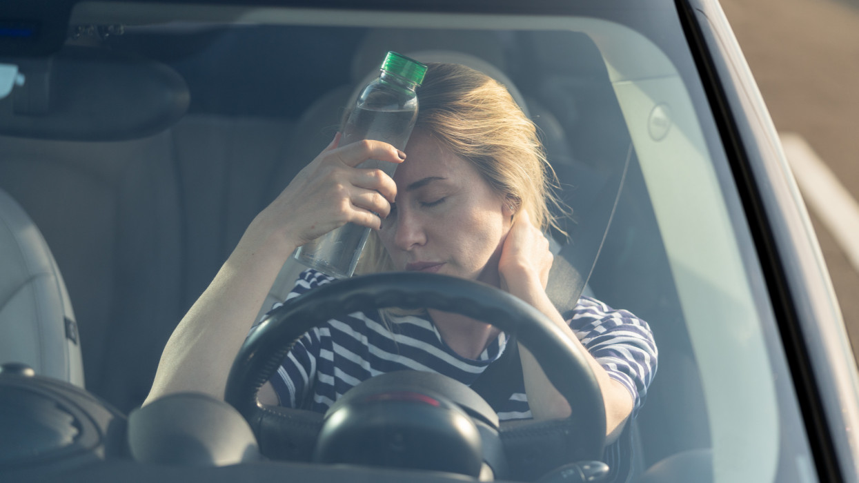 Exhausted woman driver feeling headache, sitting inside her car, applies bottle of water to forehead, hot weather. Tired female stop after driving car in traffic jam. Blood pressure, heat concept.