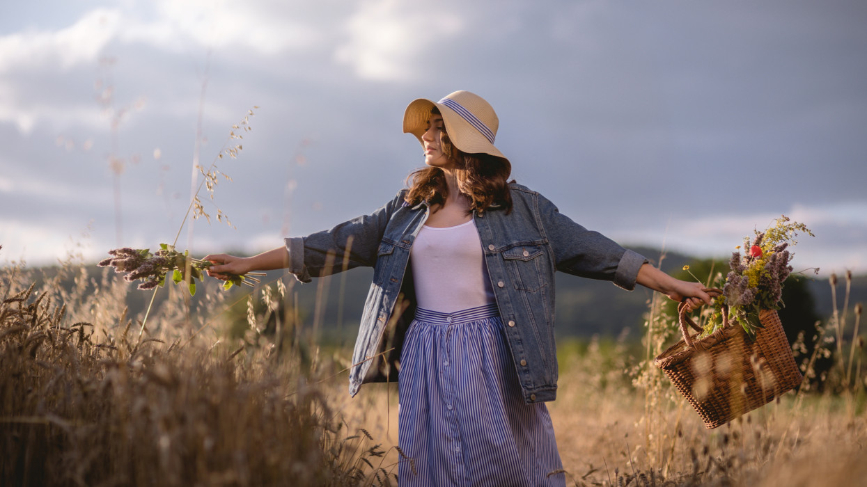 Lovely and gorgeous hipster girl with a basket full of flowers and a hat, traveling through a golden wheat field in a denim jacket.