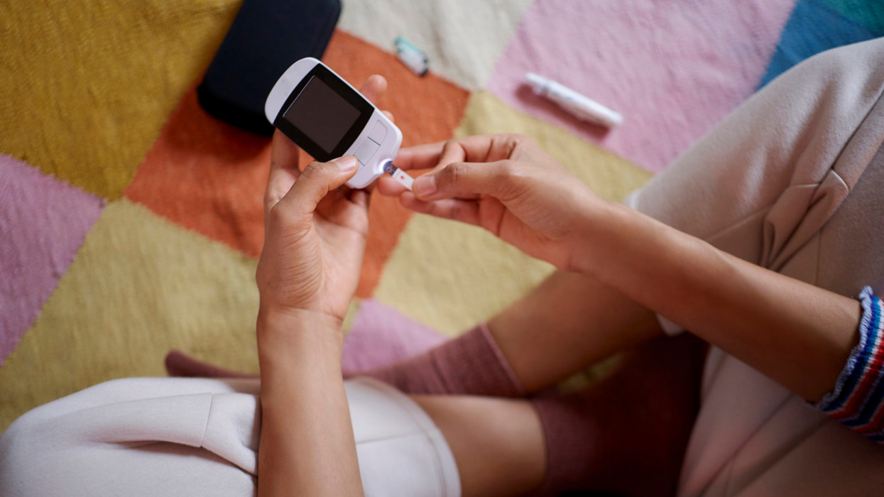 Young woman measuring her blood sugar using glucometer sitting on carpet in the living room.