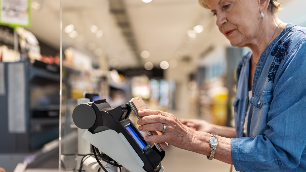 Senior woman making a mobile payment for buying groceries at the supermarket checkout. Elderly female customer paying for groceries purchase at the department store.