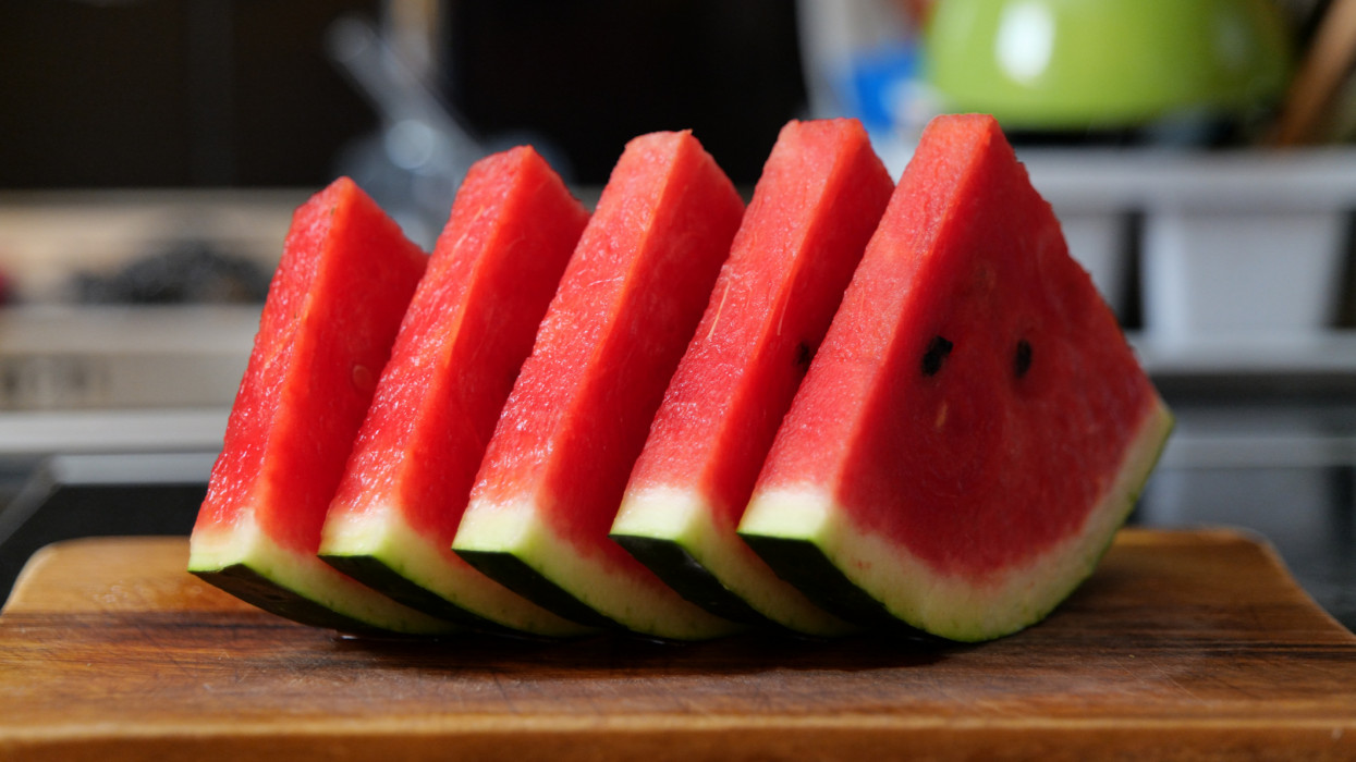 On the picture there are five slices of a watermelon lying on a wooden chopping board in the kitchen of a home. In the background we see the sink and other things related to dishwashing.