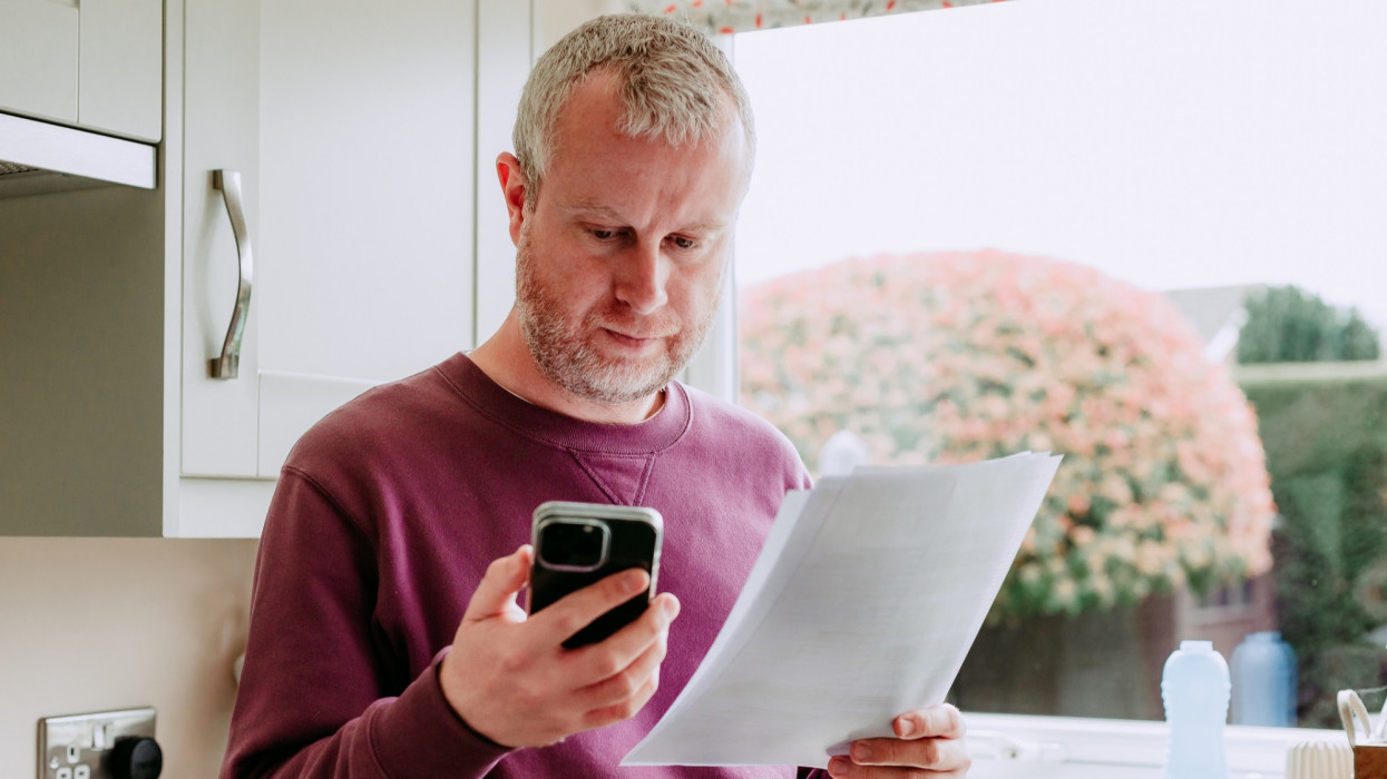 Portrait of a mid adult man in his 30s checking his energy bills at home. He is using his smart phone device to pay the bills. He is dressed casually in a purple sweater and jeans while in his modern kitchen at home.