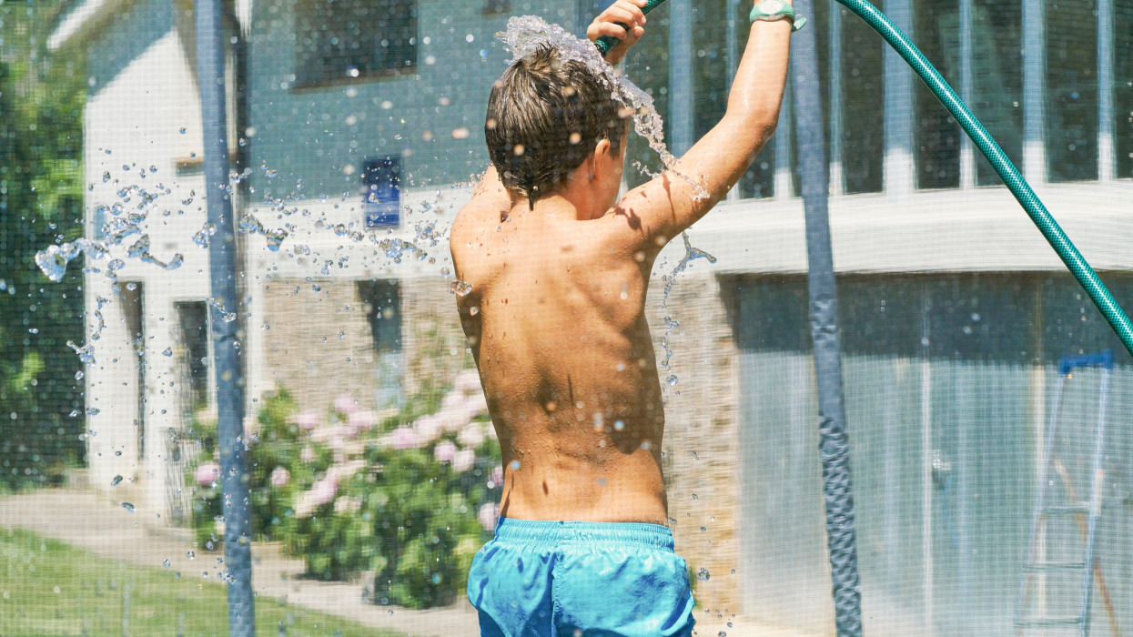 Waist up back view caucasian child getting wet with a hose in the garden of his house at summer sunset