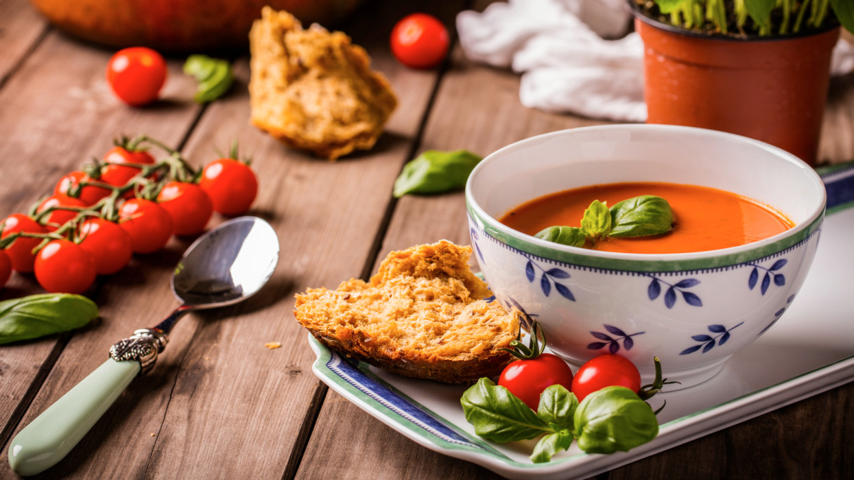 Tomato and basil soup shot on wood boards angled view with vine tomatoes loose basil spoon copper pot basil plant bread landscape