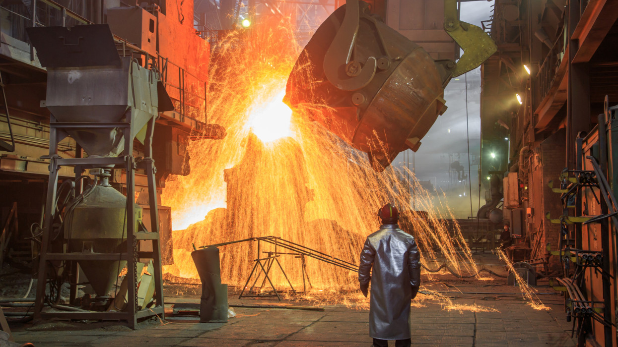 Hot steel pouring from big casting ladle into a mold in an iron foundry. Sparks, flame and smoke on the background. Factory worker controls the process.