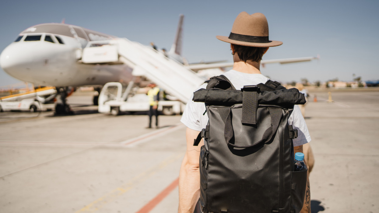 white young man with hat and suitcase walking through the airport near the takeoff runway