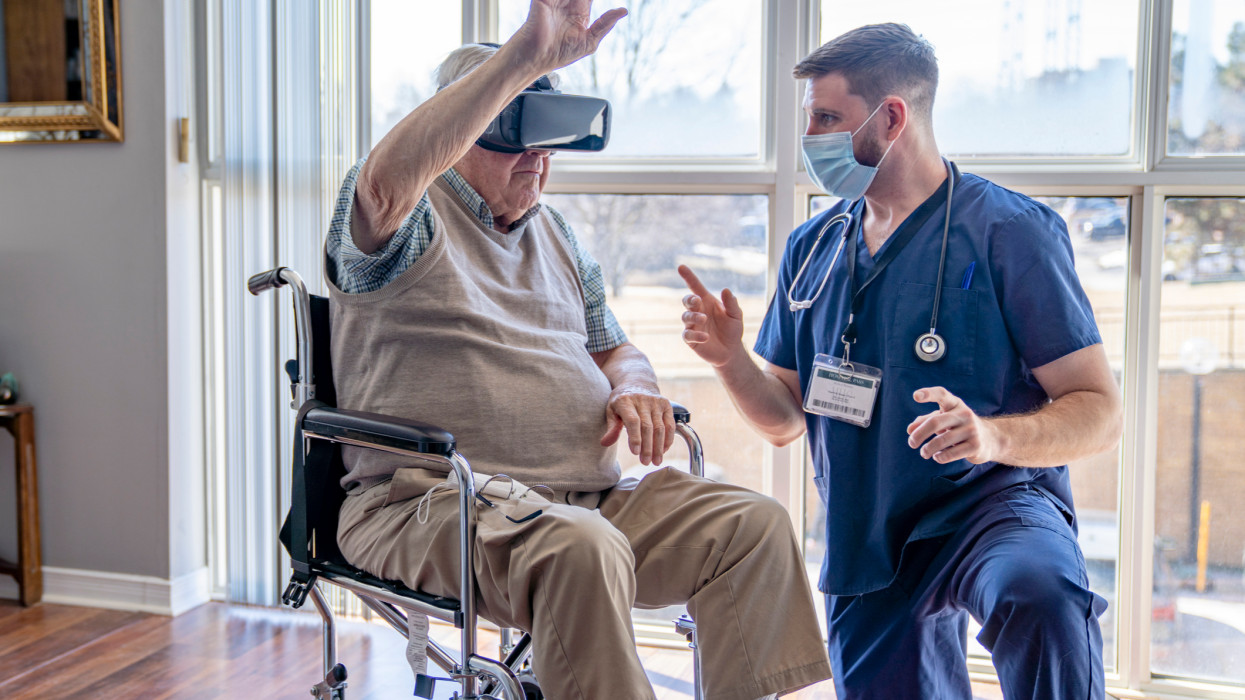 A gentleman in his 90s sitting in a wheelchair wearing a virtual reality headset and using it as apart of a new form of therapy. He overseen by a medical professional to ensure safety and proper use.