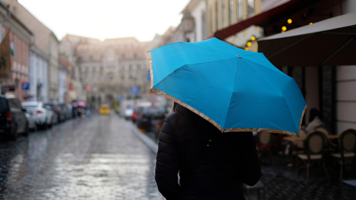 City of Budapest, tourist seen from behind walking with a blue umbrella along the street of the historic center while it is raining.