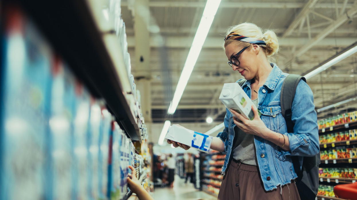 Beautiful blonde women wearing glasses, in a denim jacket and a skirt, shopping for milk in the supermarket. She is holding two cartons of milk and choosing which one to buy.