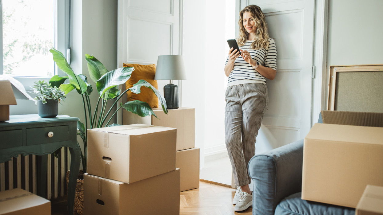 Mature woman moves in to new home, unpacking boxes and enjoying in her new home. She is resting and using smart phone.