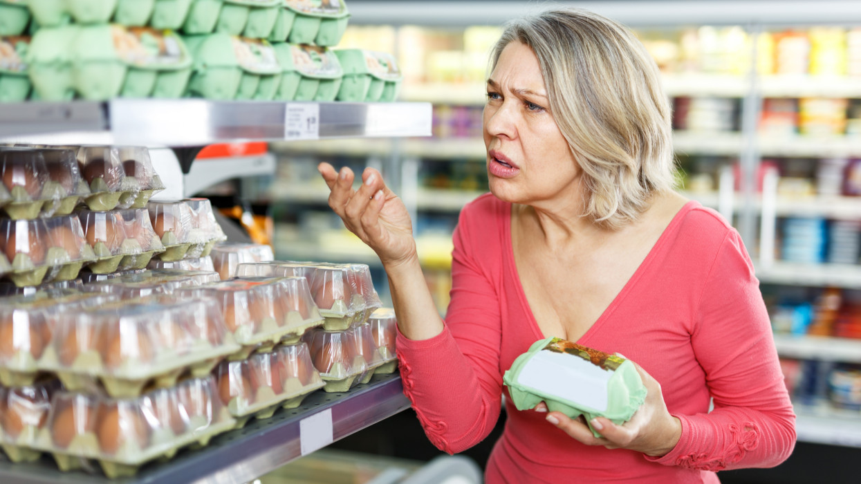 Unpleasantly surprised mature woman looking at price tag when choosing eggs in supermarket