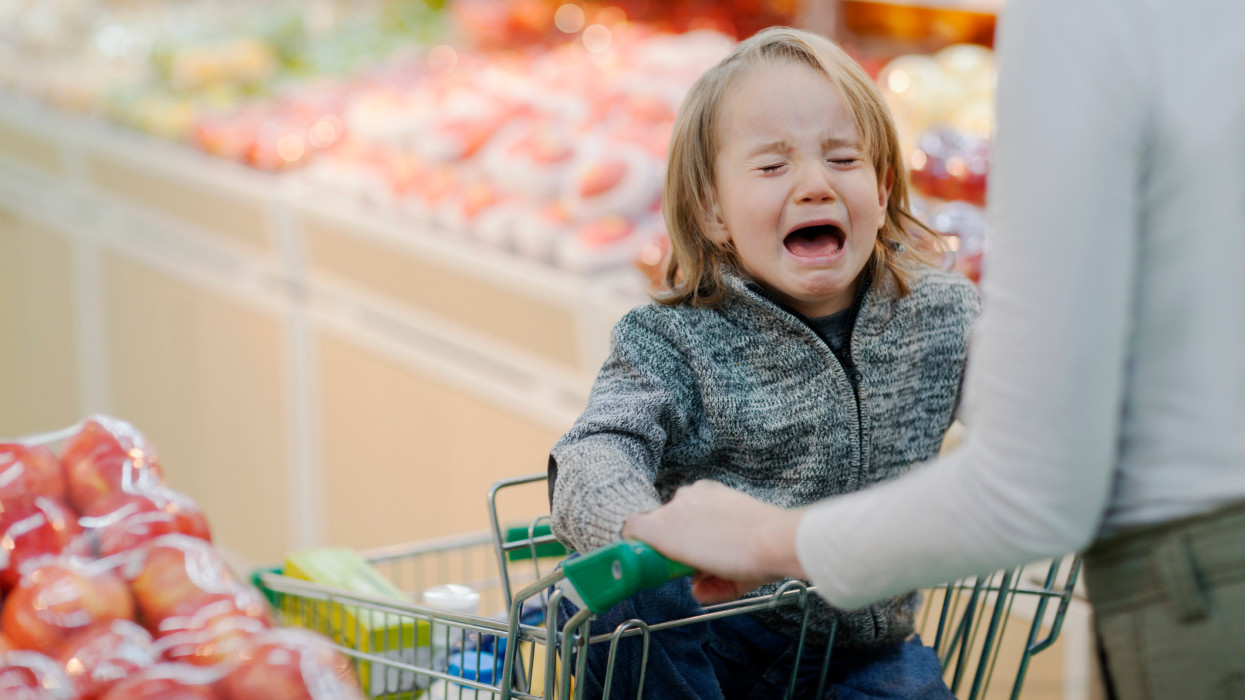 Young boy child crying temper tantrum in shopping cart with mother parent in produce section aisle of supermarket growing pains