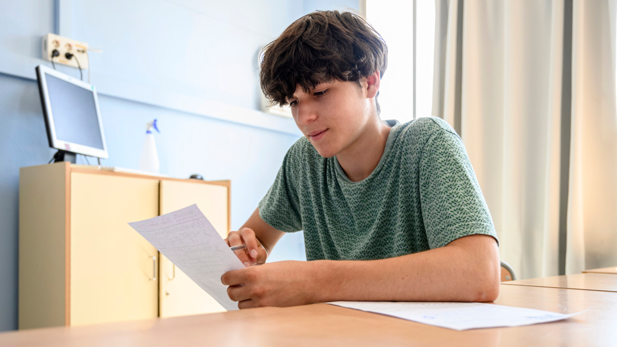 Male teenager student holding a paper for reading while sitting at classroom