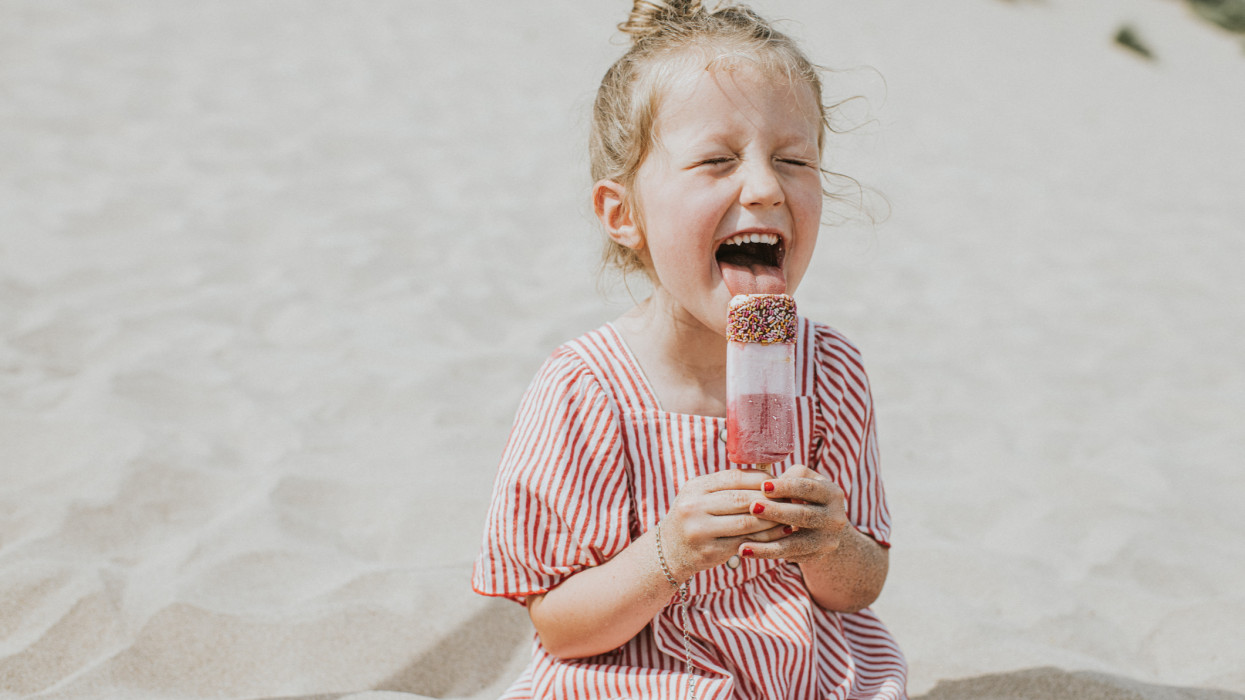 A cute young girl makes an over exaggerated expression while licking a yummy ice lolly on a summers day. She sits on a sandy beach in a little sun dress, with her hair in a top knot. She sticks out her tongue and closes her eyes. Sand provides space for copy.