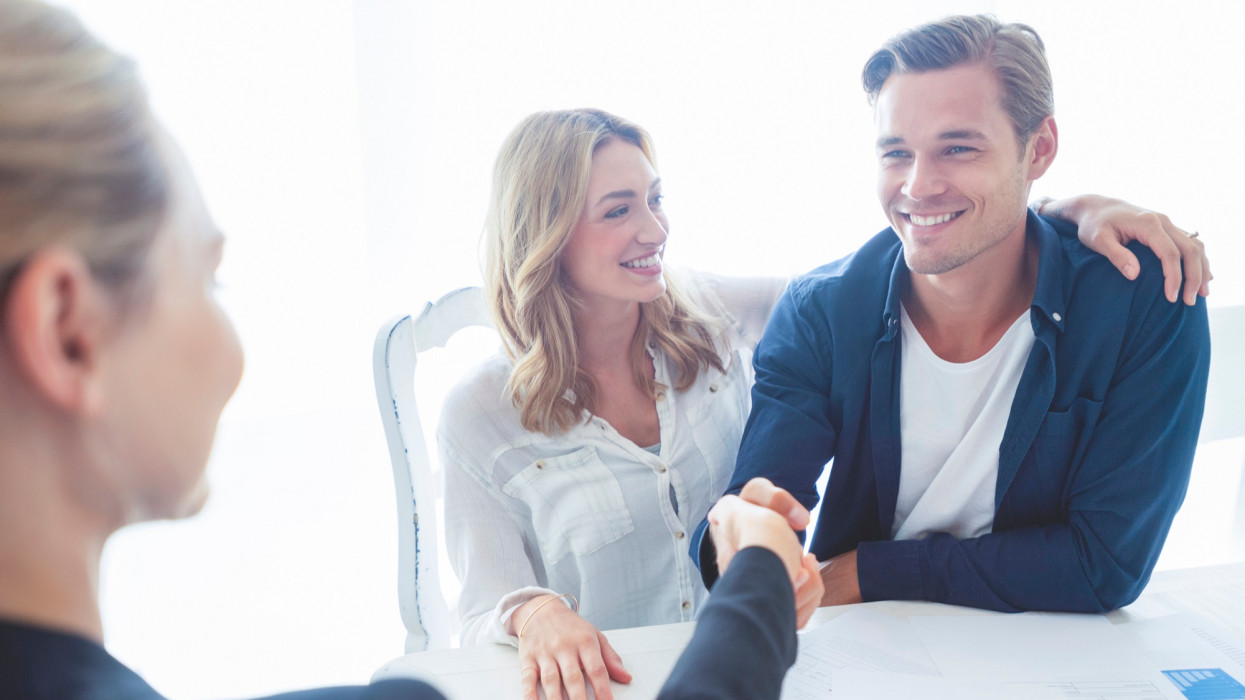 Real estate agent with couple shaking hands closing a deal. There is a digital tablet on the table. Couple are casually dressed. They are looking happy and smiling. Over the shoulder view inside a home.