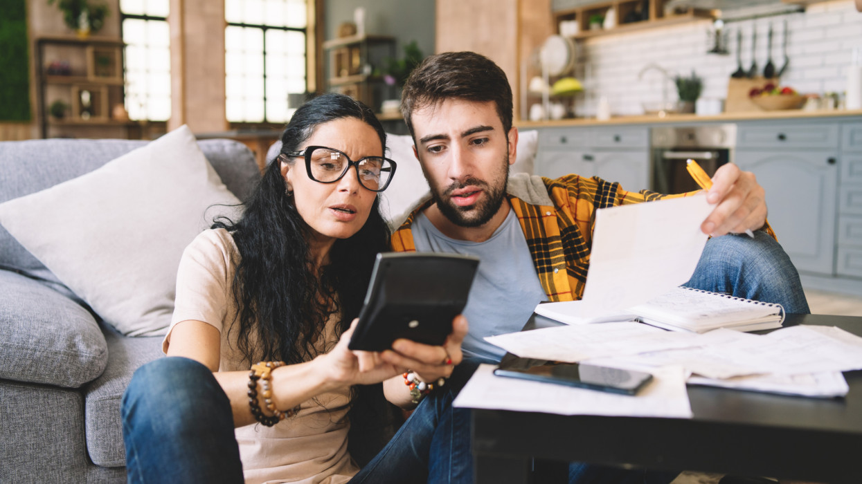 Couple calculating bills at home using calculator.  Couple checking bills and documents while calculating finances sitting in the living room. Man and woman at home analyzing their finance with documents.