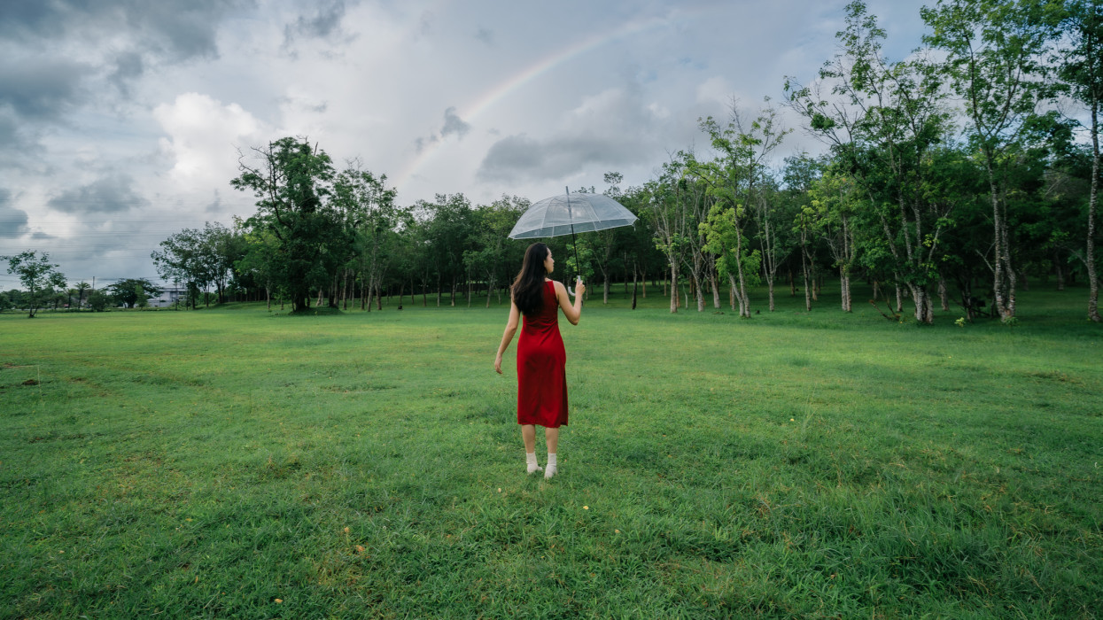 Woman in red dress spreading umbrella in rain and rainbow at green field and trees