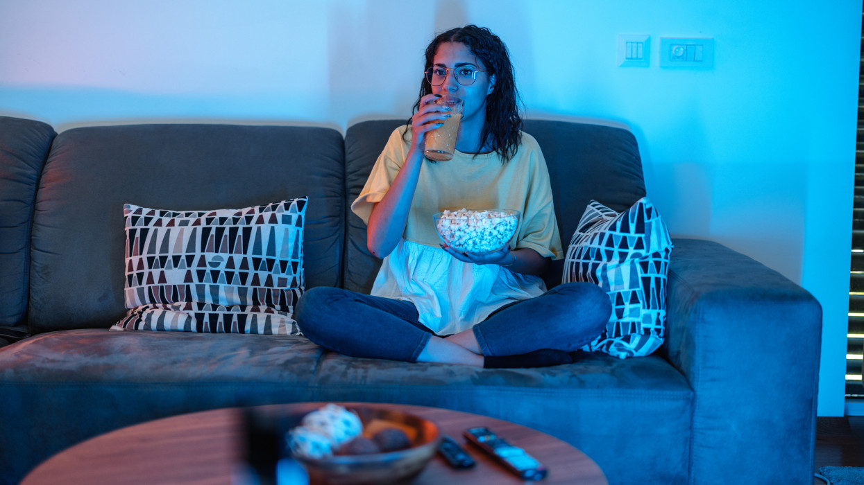 Young latin female with glasses is sitting on her sofa, drinking natural orange juice and holding popcorn while watching her favourite show.