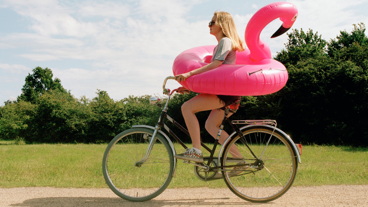 Young woman cycling though park with pink flamingo swim ring.