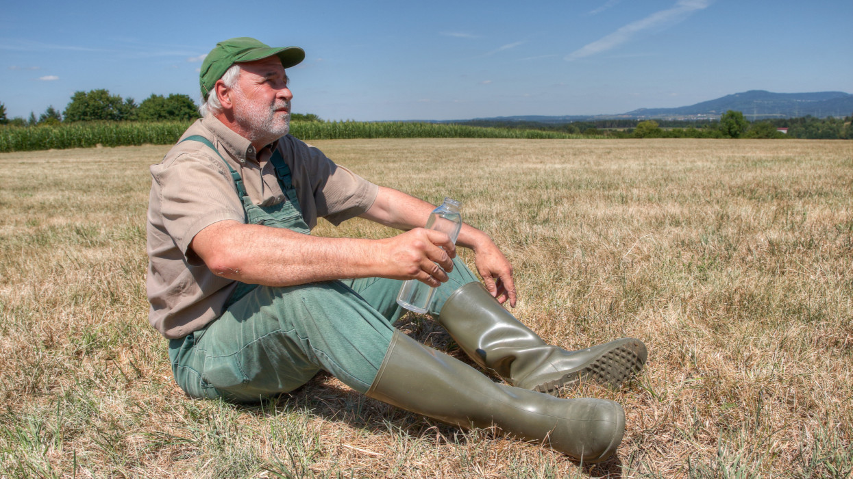 A farmer sits helplessly on his parched meadow with a bottle of water in his hand and looks thoughtfully at the sky. Climate change is causing his farmland to dry out more every year.