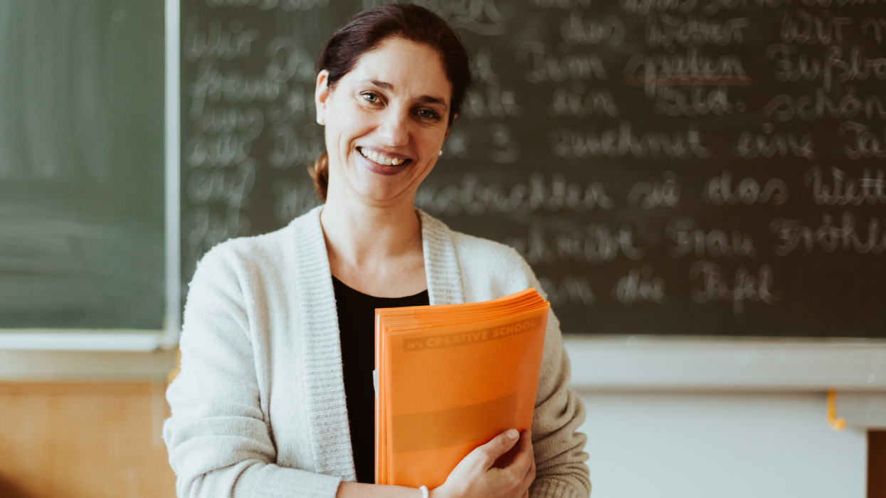 Portrait of a smiling female teacher with dark bound hair who is standing in a class room in front of a chalkboard.