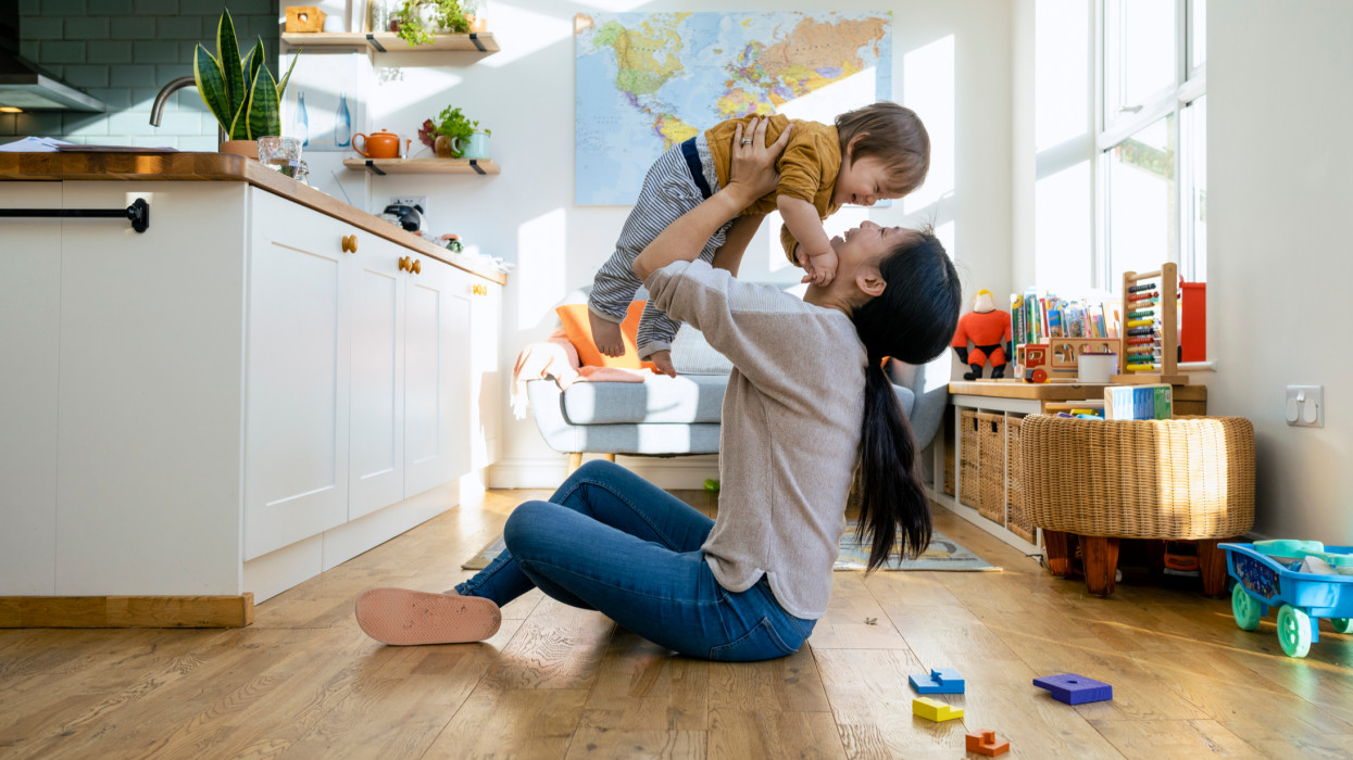 A side-view shot of a mid-adult mother sitting on the floor of a living room with her young boy she is lifting him up in the air, they are wearing casual clothing playing and laughing together.