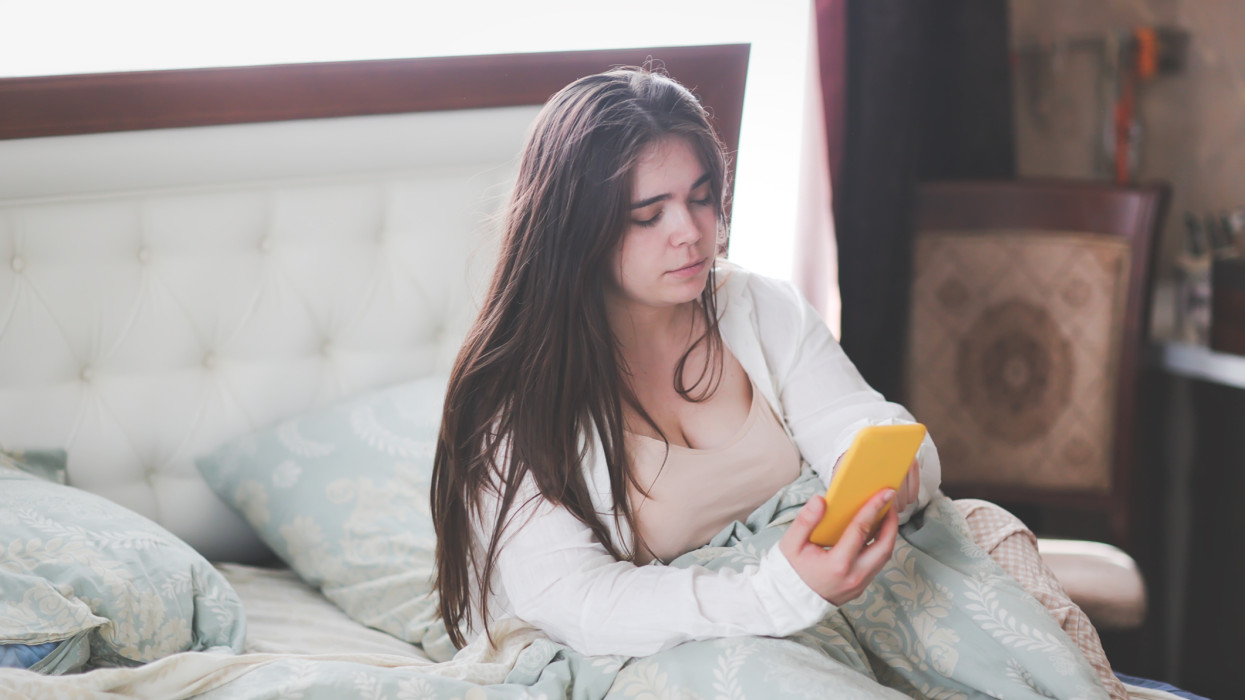 Plus size young woman in home bedroom on bed with phone.