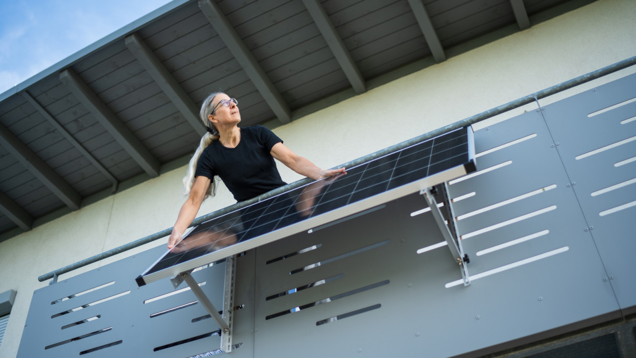 mature adult woman adjusting solar panel on her balcony in right direction to the sun, view from garden up