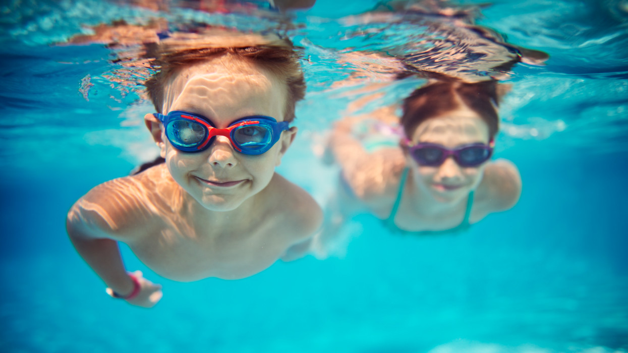 Smiling kids enjoying underwater swim in the pool towards the camera. Sunny summer day. Brother aged 5 is in the front, the sister is aged 9 and is swimming in the background. Slightly soft.