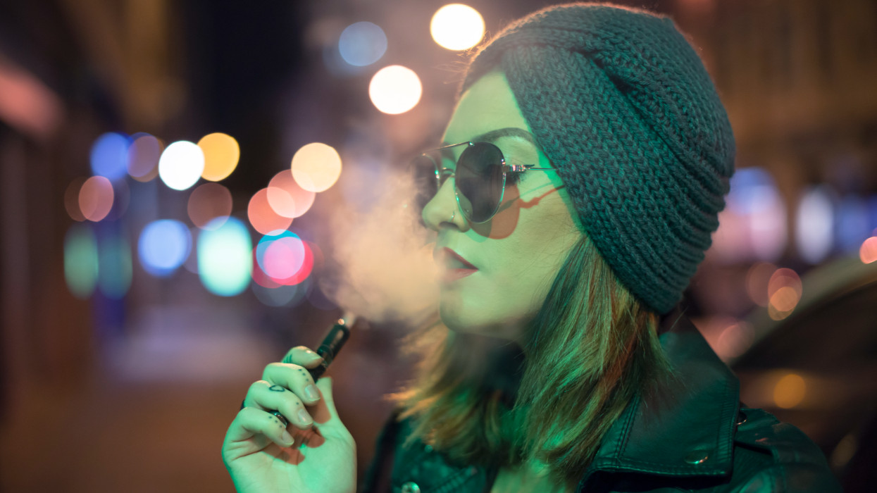 Portrait of young woman on the night street smokes electronic cigarette.