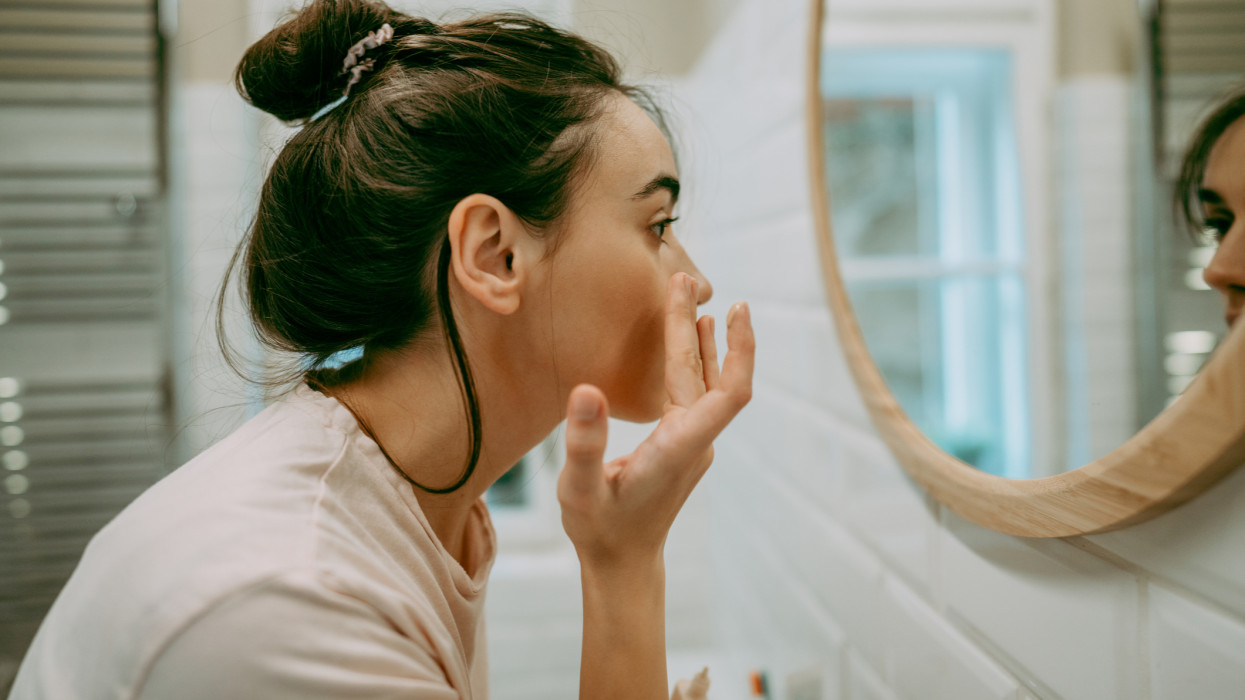 Photo of a young woman getting ready in the bathroom for the upcoming day; applying moisturizer as a part of her daily routine.