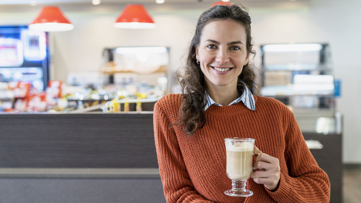 Smiling young woman holding coffee glass mug while standing inside filling station store during daytime