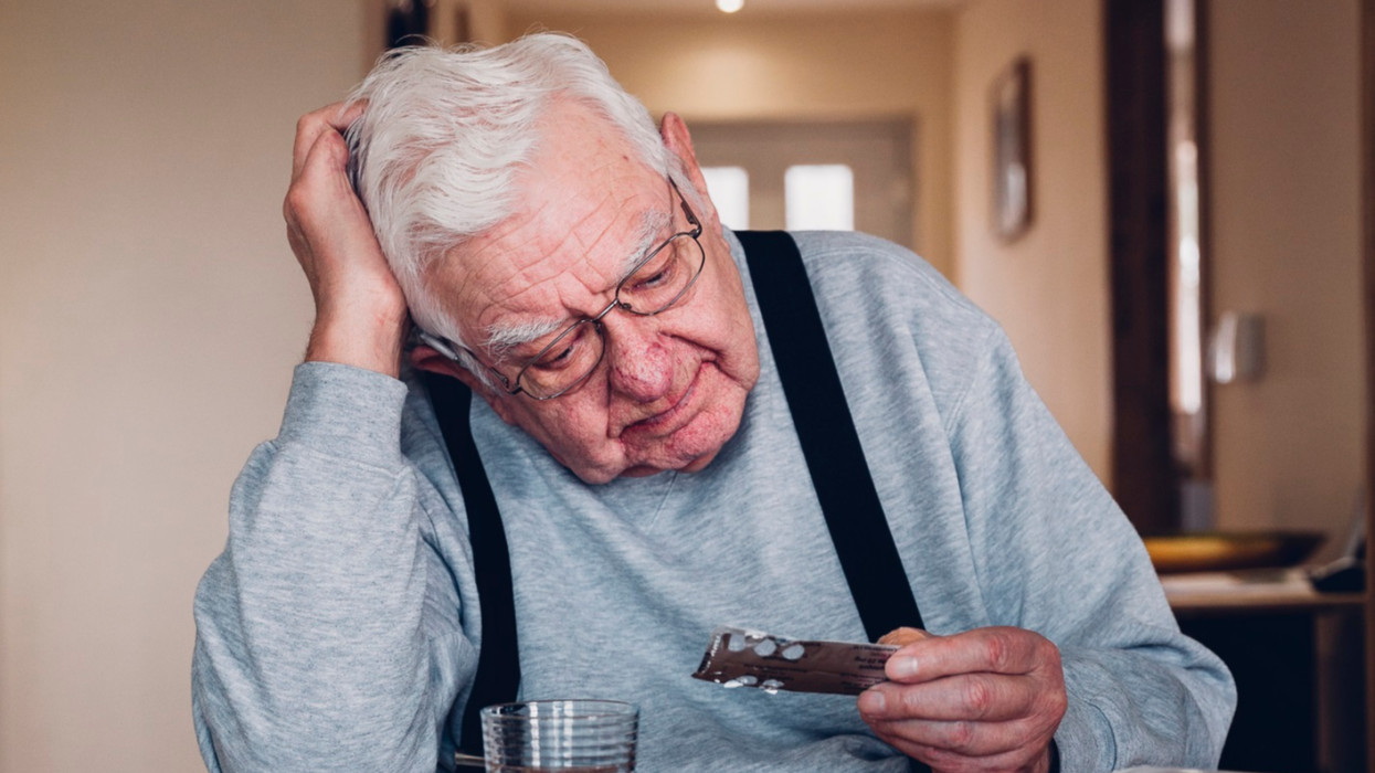 Elderly man sitting at home at a table with his head resting on his hand. Looking troubled he holds a packet of pills with other medication around him