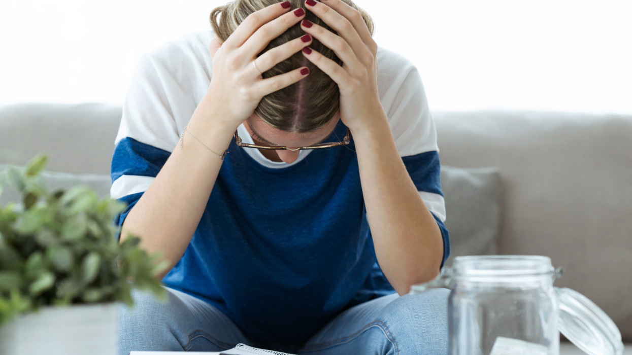 Shot of depressed young woman holding her head with hands while counting savings at home.