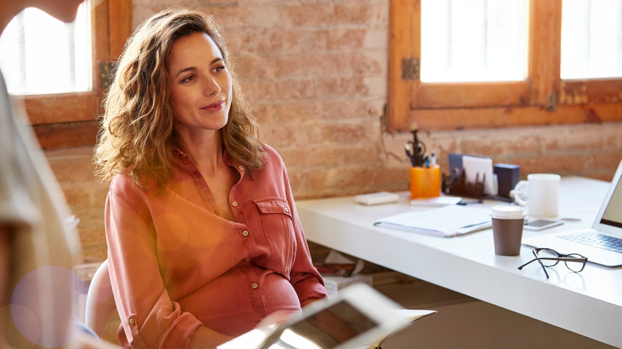 Pregnant businesswoman looking away at desk. Beautiful female professional is sitting in office. She is wearing shirt at workplace.