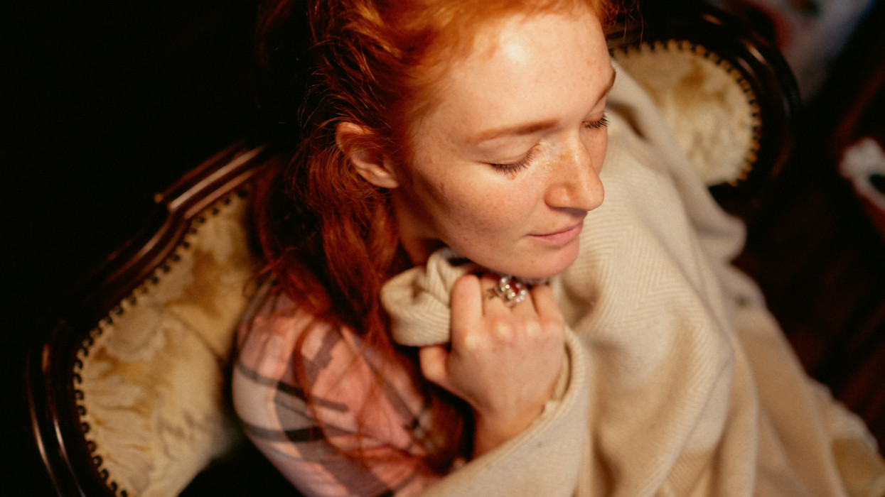 Portrait of a young red-haired woman. She is sitting in an armchair and has wrapped herself in a warming blanket. She has her eyes closed.