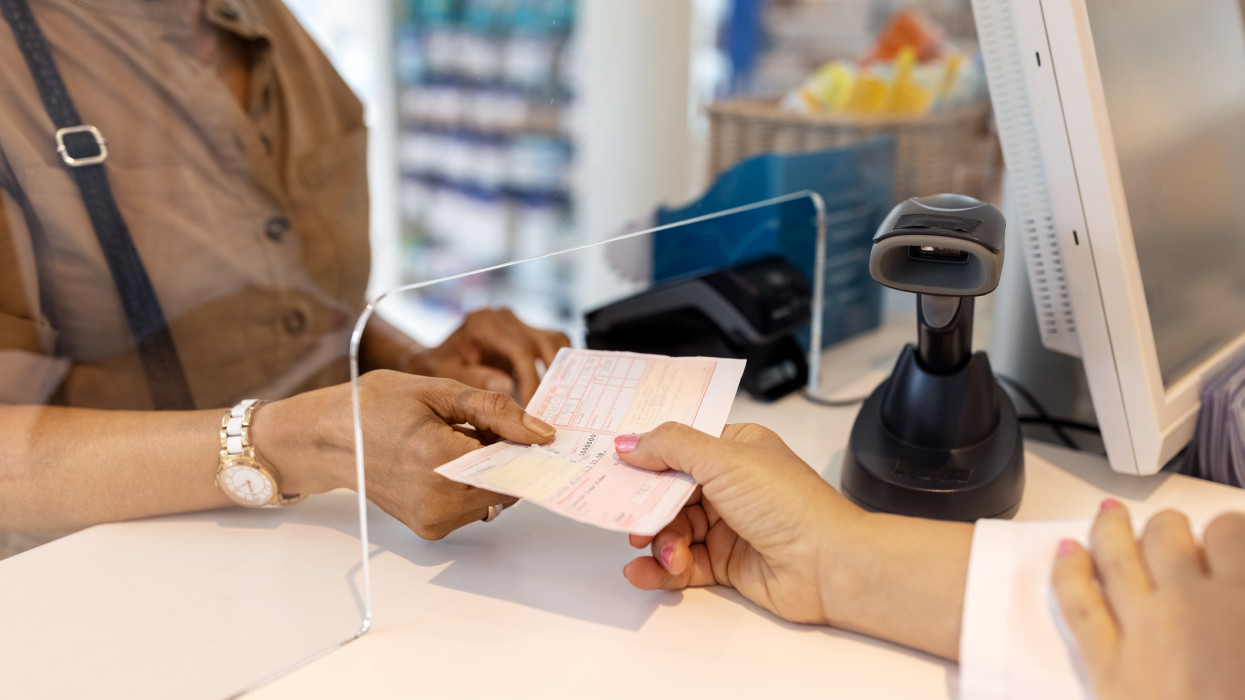 Cropped image of woman giving the medicine prescription to female pharmacist in store. Close-up of chemist hand taking prescription paper from customers hand at the pharmacy.