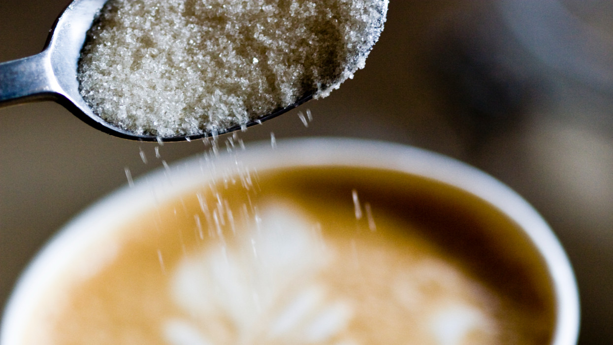 A spoon of sugar being poured into a latte.  Latte has a pattern with milk and espresso.