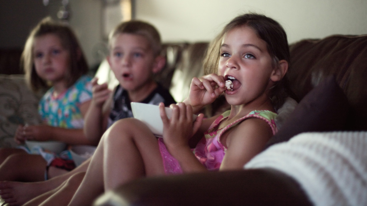 Children seated on leather couch, eating popcorn watching movie.