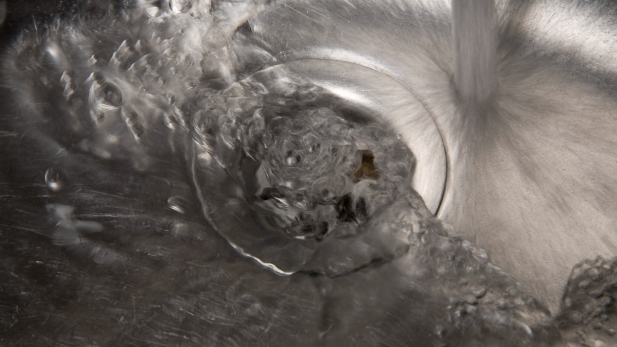 DSLR photo of moving, swirling water, coming from a kitchen tap, starting to underline the counter-clockwise Coriolis effect, streaming via a drain in a stainless steel sink to disappear in the discharge pipeline system. The sink is found in domestic but also commercial kitchens. The stainless steel looks black and white, but is photographed in color. Water takes every form.