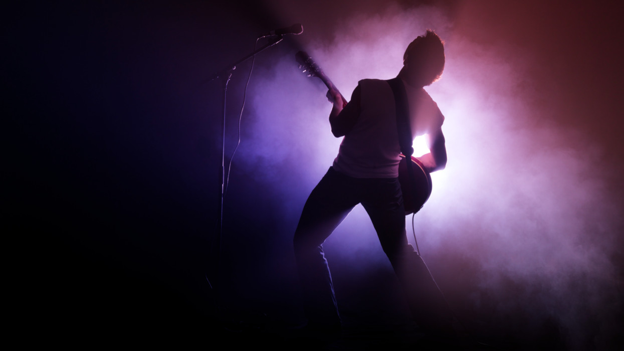 A male guitarist performs on stage surrounded by strong light and smoke. Full body shot photographed from behind.